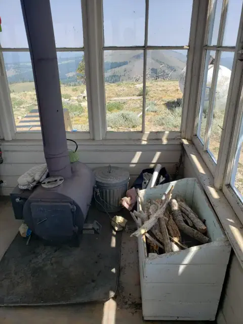 The interior of a small cabin features a wood-burning stove and a box of firewood.
