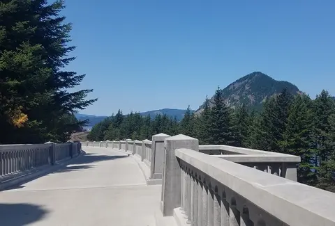 A concrete bridge curves through a landscape of green trees and distant mountains under a blue sky.