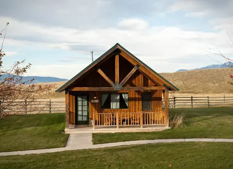 A wooden cabin stands in a grassy field with a fence and mountains in the background.