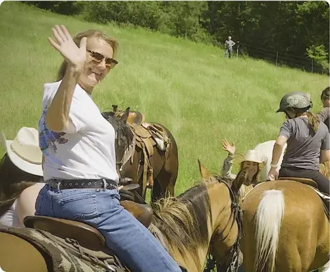 A woman waves while riding a horse in a grassy field.