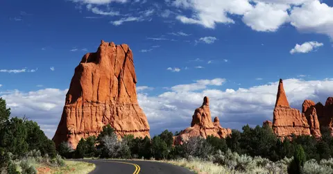 A paved road curving through a landscape with red rock formations and a blue sky with clouds.