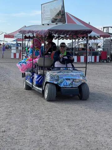 A decorated golf cart carrying people and a clown drives outside near tents.