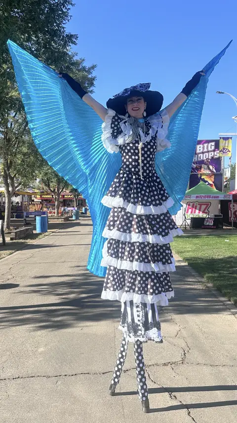 A performer on stilts wears a polka dot dress and large blue wings.