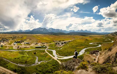 A hiker stands on a rocky overlook looking out over a wide valley with a winding river and distant mountains.