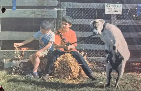 Two boys sitting on hay bales next to a standing goat.