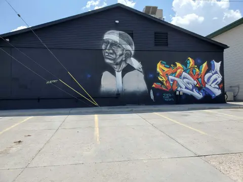 A black building features a large mural of an Indigenous person alongside colorful graffiti.