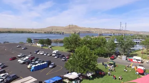 An aerial view shows a parking lot and tents set up near a river with a large bridge in the background.