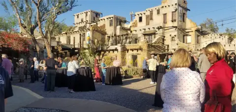 A gathering of people standing around cocktail tables in front of a historic adobe building exterior.