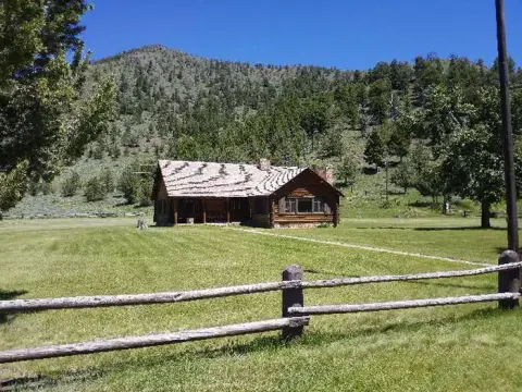 A wooden cabin sits in a grassy field with a mountain in the background.