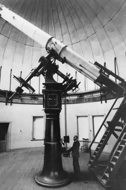 Black and white photo of a large telescope inside an observatory dome with a person standing next to it.