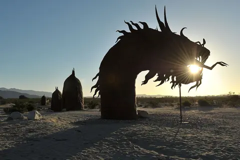 A large metal serpent sculpture stands in a desert landscape with the sun shining through its mouth.