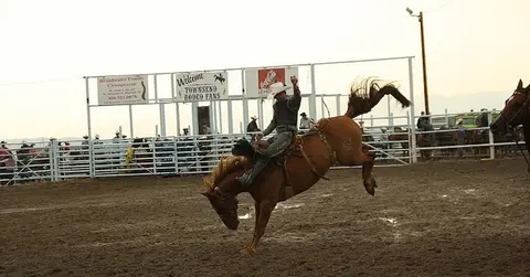 A cowboy rides a bucking horse at a rodeo arena.