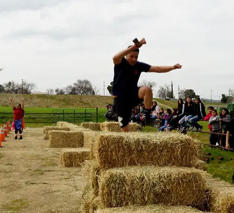 A young person jumps over stacked hay bales outdoors while a crowd watches.