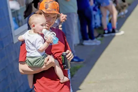 A person carrying a baby who is drinking from a bottle outdoors.