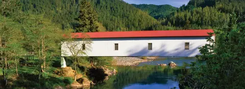 A white covered bridge with a red roof crosses a calm river surrounded by lush green forests and hills.
