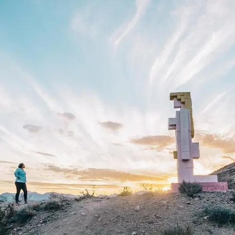 A person stands looking at a large abstract sculpture outdoors at sunset.