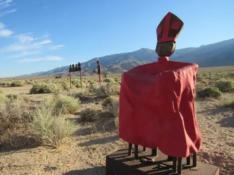 A large red sculpture stands in a desert landscape with mountains in the background.