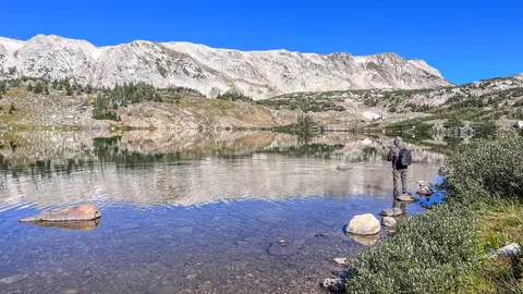A person fishes in a clear alpine lake below a rocky mountain ridge.