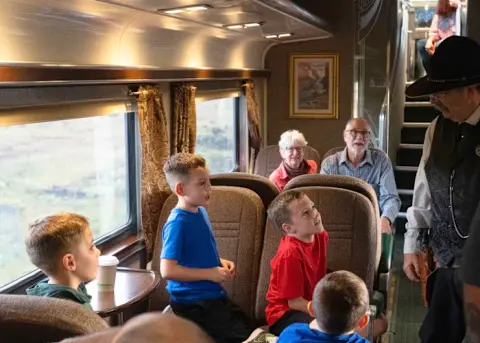 Passengers are seated inside a train car as a man in a hat talks to them.