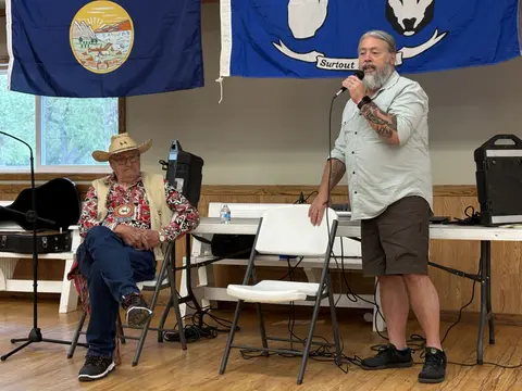 A man speaks into a microphone while another man sits nearby indoors.
