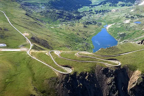 An aerial view shows a winding mountain road near a blue alpine lake.