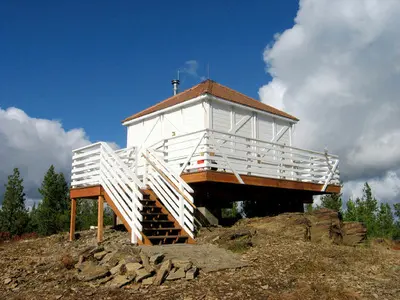 A white cabin with a viewing deck sits atop a rocky hill under a blue sky.