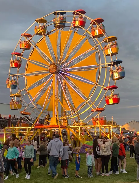 A large ferris wheel glows with yellow and colored lights at an evening fair or carnival with a crowd of people below.