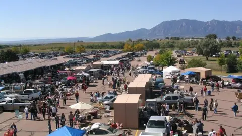 An outdoor flea market with many stalls and parked cars under a bright sky with mountains in the distance.