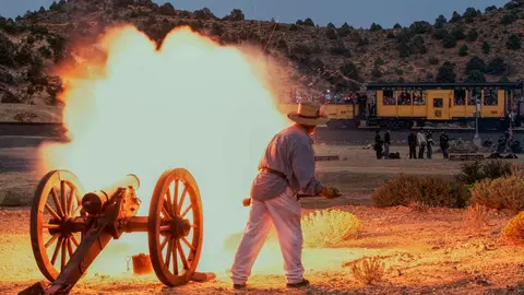 A person fires a cannon with a large burst of smoke and fire in front of a train.