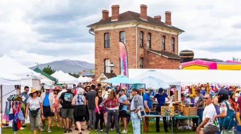 A busy outdoor street market with many vendors, tents, and shoppers in front of a historic brick building.