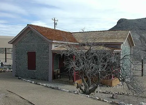 A small building with walls constructed from glass bottles stands in a desert landscape.