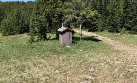 A small wooden outhouse stands in a clearing near a dirt path and a forest.