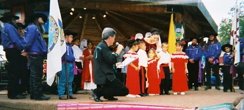 A man speaks into a microphone on a stage while children and adults in cultural attire stand around him.