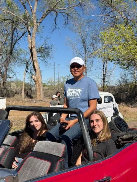 Three people pose in a red convertible car outdoors.