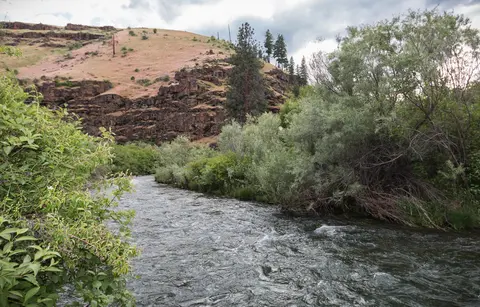 A swift river flows past a grassy bank and a rocky hill.
