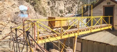 An old mining cart sits on a yellow trestle track outside a corrugated metal building against a rocky hillside.
