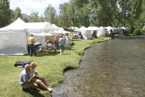An outdoor market with white tents set up along a grassy riverbank.
