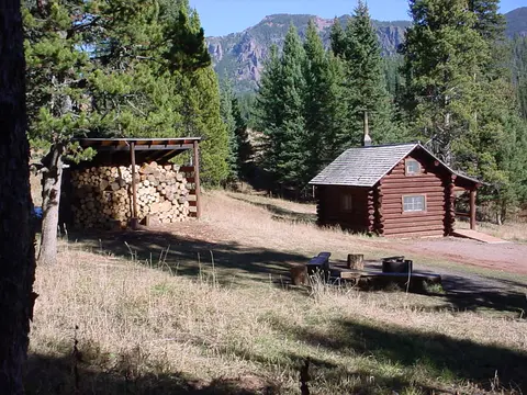 A log cabin and a wood shed sit in a mountain clearing surrounded by trees.