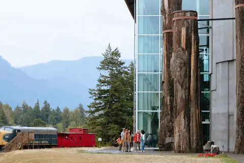 People walk past carved wooden pillars outside a glass building with mountains and a train in the distance.