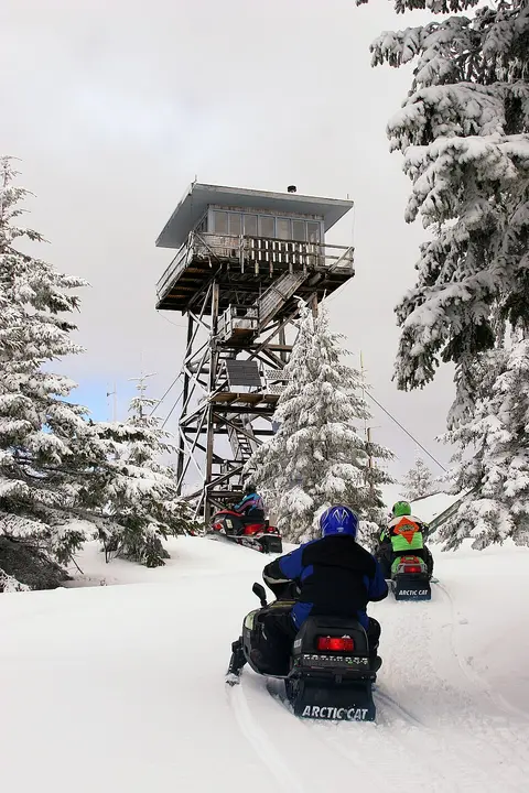 People ride snowmobiles on a snowy trail near a fire lookout tower.