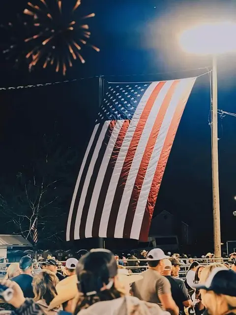 A large American flag hangs at night over a crowd watching fireworks.
