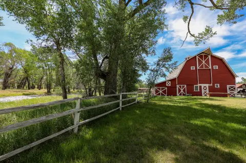 A red barn sits near a wooden fence and large trees under a blue sky.