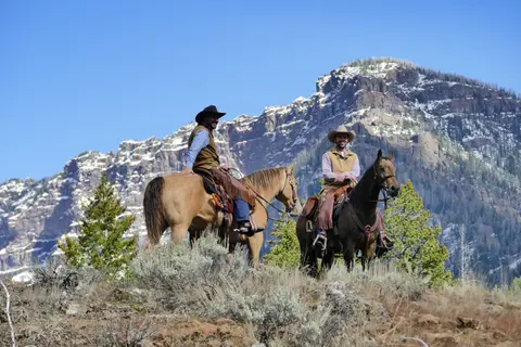 Two cowboys on horseback pose in a landscape with mountains in the background.