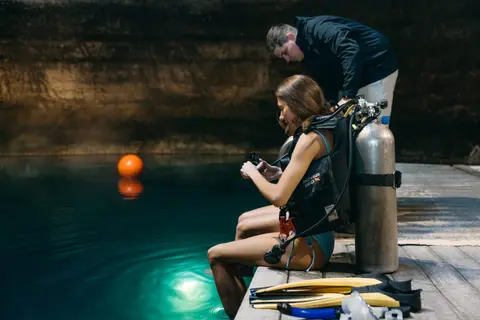 A woman prepares to dive into the water with scuba gear while a man assists.
