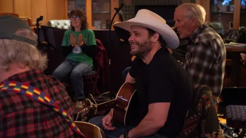 A man wearing a cowboy hat plays a guitar indoors with a group of people.