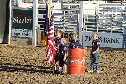 Four children in scout uniforms holding flags in an outdoor arena.