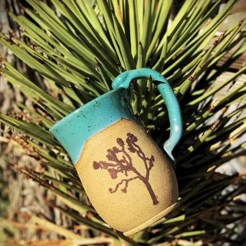 A pottery mug with a turquoise glaze and a tree design rests against green foliage.