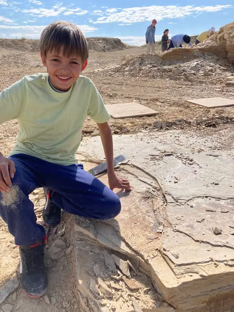 A smiling boy kneels by a fossil embedded in a rocky excavation site.