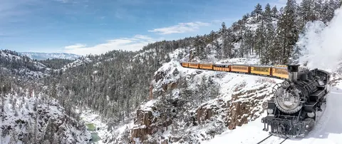 A steam train travels along a snowy mountain ledge above a canyon.