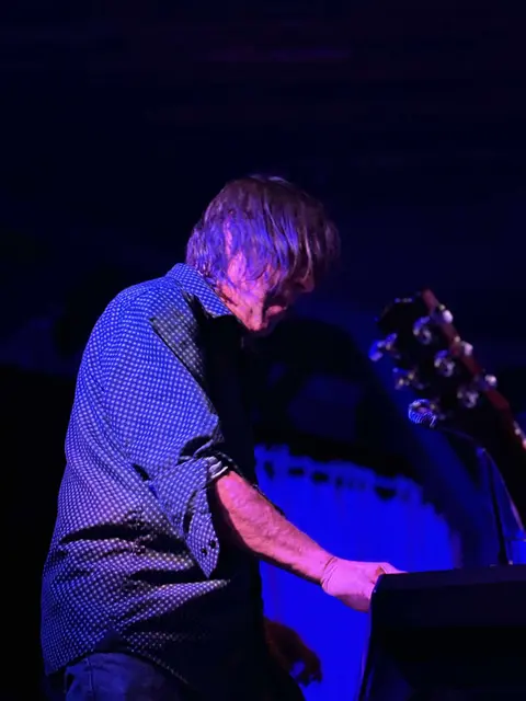 A man plays a keyboard on a stage under blue lighting.